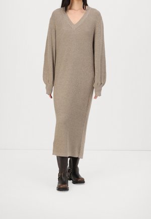 Mujer con un vestido largo de punto acanalado beige con escote en V y mangas abullonadas combinado con botas de cuero negras hasta media pantorrilla con hebillas.