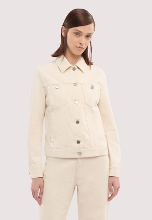 Donna con capelli castani lisci che indossa una giacca di jeans color crema con bottoni e pantaloni abbinati, in piedi davanti a uno sfondo semplice.