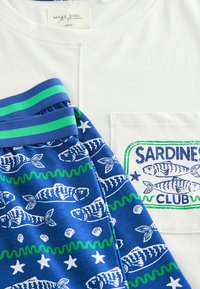 White cotton t-shirt with a pocket featuring "SARDINES CLUB" print. Blue shorts with white fish and starfish patterns, green striped waistband.