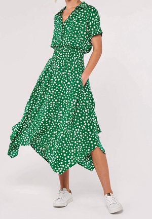 Robe verte à pois blancs, manches courtes, col à boutons, taille élastique, jupe fluide et poches latérales. Portée avec des baskets blanches.