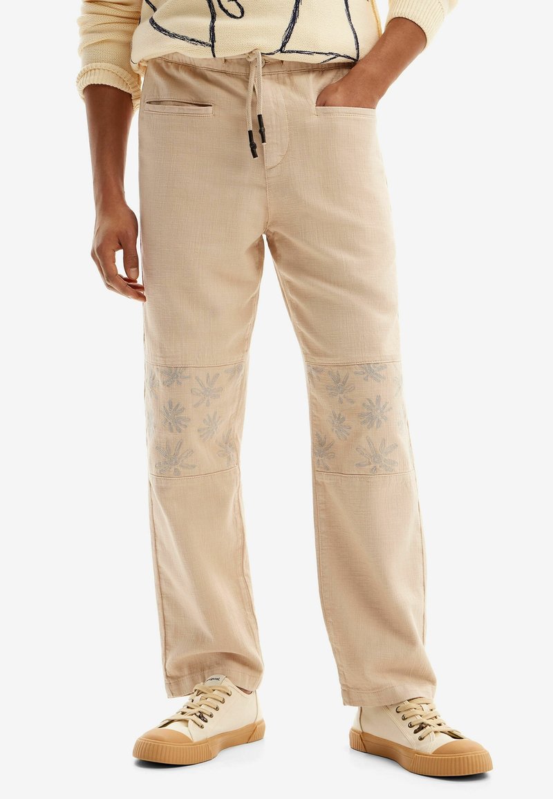 Pantalon beige à cordon avec des patchs floraux aux genoux, porté avec des baskets crème et un pull léger avec des lignes noires abstraites.