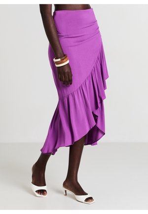 Femme portant une jupe asymétrique à volants violette, des talons blancs à bouts ouverts, et des bracelets en bois et crème au poignet, debout sur un sol clair.