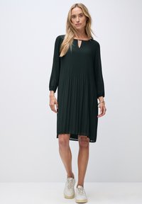 Dunkelgrünes, plissiertes Kleid mit langen Ärmeln und einem Schlüsselloch-Ausschnitt. Getragen mit weißen Sneakers, mit lockerer Passform und asymmetrischem Saum.