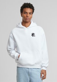 Haut à capuche blanc en tissu doux avec une poche kangourou et des poignets côtelés. Présente un petit logo noir sur la poitrine. Coupe classique.