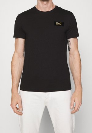 Mann trägt ein schwarzes kurzärmliges T-Shirt mit einem goldenen "EA7 Emporio Armani"-Logo auf der Brust und weiße Hose.
