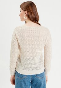 Pull en maille beige clair, avec un motif texturé et des poignets côtelés. Le dos est en maille ajourée avec une coupe décontractée sur un jean bleu.