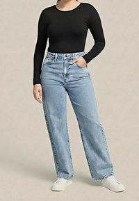 Personne portant un t-shirt à manches longues noir, un jean taille haute bleu clair à larges jambes et des baskets blanches, debout avec une main dans la poche.
