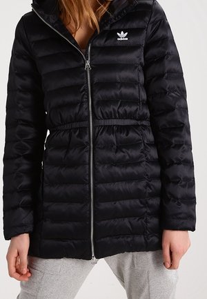 Schwarze, gesteppte Adidas-Jacke mit Frontreißverschluss, getragen von einer Person in grauer Hose, teilweise sichtbarer Oberkörper und Hände.