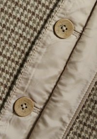 Knap, der har et struktureret houndstooth mønster i brun og beige, med et glat, beige stof og en rund, fire-huls knapdetalje.