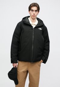 Zwarte geïsoleerde jas met capuchon, ritssluiting aan de voorkant en logo op de borst. Het model draagt een lichtbruine broek en houdt een zwarte pet vast.