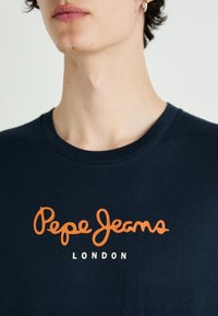 Tamsiai mėlynas medvilninis marškinėlis su oranžine užrašu "Pepe Jeans" ir mažesniu baltu tekstu "LONDON" apačioje. Apvalaus kaklo dizainas.