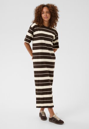 Femme aux cheveux bouclés debout, portant une robe longue ample à rayures marron et crème à manches courtes et des baskets marron, les mains derrière le dos.