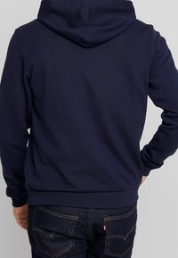 Námořnická modrá mikina s kapucí na šňůrku, dlouhými rukávy a žebrovaným pasem. Nosit s tmavými džíny, ukazující minimalistický design.
