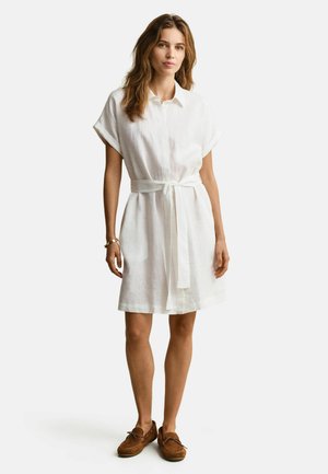 Femme debout portant une robe chemise blanche à manches courtes avec ceinture et des mocassins marron sur un fond clair uni.
