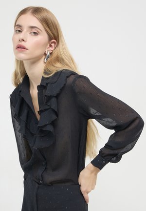 Eine Frau mit langen blonden Haaren trägt eine transparente schwarze Bluse mit Rüschen und kleinen strukturierten Punkten sowie großen silbernen Creolen.