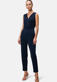 Navy blue jumpsuit with a V-neck, sleeveless design, pleated waist, and tapered legs. Made of smooth material; paired with strappy heels.