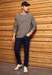 Šedý texturovaný svetr, tmavé džíny a bílé tenisky s černými akcenty. Osoba má na sobě námořnickou čepici a stojí proti dřevěným obkladům.