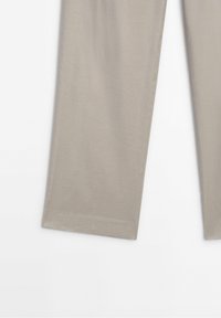 Partie inférieure d’un pantalon beige droit posé à plat sur une surface blanche, montrant les détails de l’ourlet et de la texture du tissu.