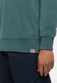 Mörkgrön sweatshirt med ribbade ärmslut och nederkant. Har en liten Dickies-logotyp sydd vid nederkanten. Slät tygkvalitet.