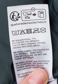 Vårdguide för ett plagg, med vit tyg och svart text. Innehåller tvättanvisningar och sammansättning: 77% polyester, 23% spandex.