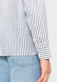 Camicetta a righe in tessuto leggero con righe orizzontali blu navy e bianche, dettagli ai polsini con bottoni, abbinata a jeans blu chiaro.
