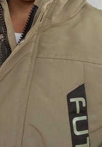 Khaki jacket featuring a zippered front and hood, with a large black label displaying "FUT." Textured fabric with a smooth finish.