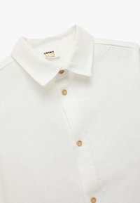 Camisa de algodón blanca con cuello tipo camiseta, que cuenta con cinco botones de madera y una superficie de tela texturizada. La etiqueta dice "Koton KIDS."