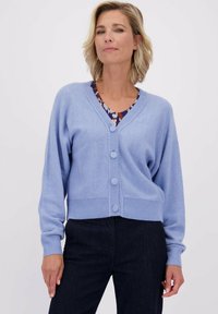 Lichtblauwe cardigan van zacht materiaal, met een V-hals, geribbelde manchetten en vijf grote knopen aan de voorkant. Gecombineerd met donkere broek.