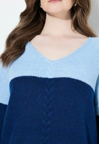 Frau trägt einen blauen V-Ausschnitt-Strickpullover mit hellem blauen oberen Bereich und dunklem blauen unteren Bereich, der detaillierte Strickmuster zeigt.