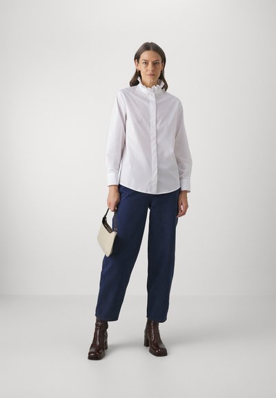 Frau in einer Bluse mit weißem Rüschensaum, dunklen blauen Hosen, dunklen braunen Stiefeln, die eine kleine beigefarbene Handtasche vor einem schlichten Hintergrund hält.