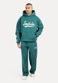 Teal fleece hoodie met een voorzak en wit logo, gecombineerd met bijpassende joggingsbroek. Eenvoudige textuur, comfortabele pasvorm. Casual sportkleding.