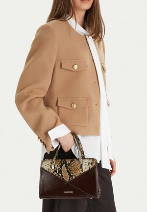 Žena v kamelovém kabátě a bílé blůze drží tmavě hnědou kabelku Lasocki s hadím vzorem.