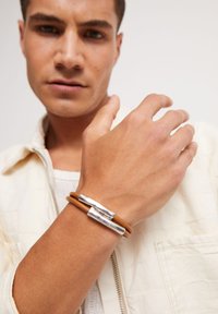 Pulsera de cuero marrón con dos detalles de metal plateado, que muestra un diseño elegante. Lleva puesta en la muñeca de un hombre con un atuendo neutro.