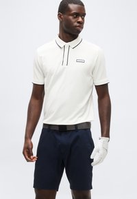 Άνδρας που φοράει λευκό πόλο με κοντά μανίκια και μαύρο περίγραμμα, ναυτικά σορτς, μαύρη ζώνη και λευκό γάντι γκολφ στο αριστερό του χέρι.