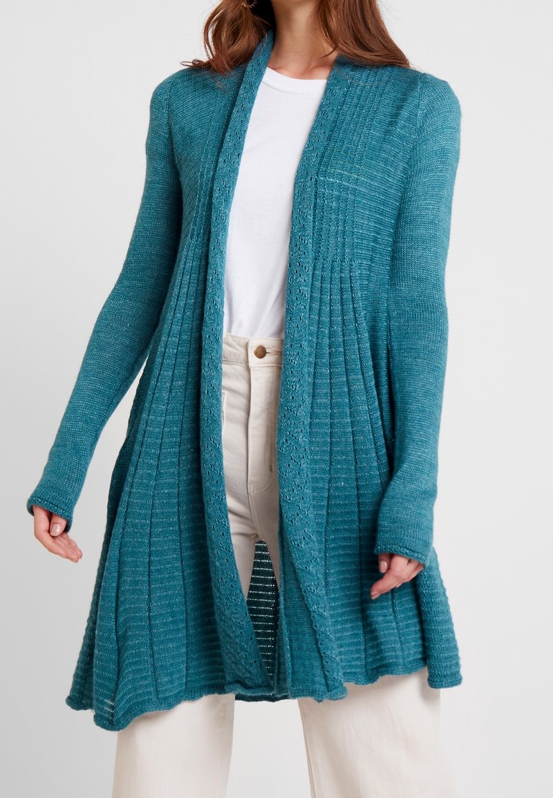 Femme portant un long cardigan tricoté couleur sarcelle avec une texture côtelée sur une chemise blanche et un pantalon beige clair, debout devant un fond uni.