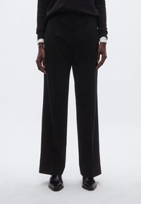 Zwarte broek met wijde pijpen en plooien, gemaakt van een gladde stof. Draagt een zwarte trui en stijlvolle zwarte schoenen.