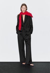 Kvinde iført en oversized sort dragt med vide bukser og et rødt tørklæde draperet over skuldrene, stående foran en ensfarvet baggrund.