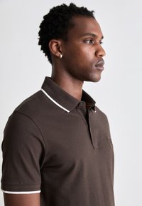 Braunes Poloshirt mit einem weißen Kragenbesatz, ausgestattet mit einer Knopfleiste und einer kleinen Brusttasche. Weicher, texturierter Baumwollstoff.