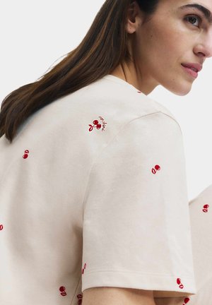 Woman with long brown hair wearing a light beige shirt with small red cherry patterns and a partial side profile of her face visible.