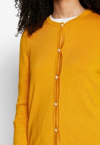 Gul kofta med rund halsringning, med knappstängning framtill och rundade, pärlliknande knappar samt ett mjukt, texturerat stickat tyg.