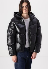 Sort puffjakke med en glat finish, quiltet design, hætte og frontlynlås. Båret med en hvid skjorte og mørke jeans.