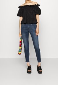 Schwarzes, schulterfreies Oberteil mit Rüschendetails, kombiniert mit dunkelblauen Skinny-Jeans und schwarzen Plateausandalen. Hält eine mehrfarbige Häkelhandtasche.