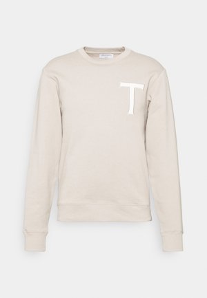 Beiger langärmliger Pullover mit Rundhalsausschnitt, gerippten Bündchen und großem weißem Buchstaben "T" auf der linken Brust.