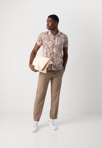 Kortærmet mønsteret skjorte med geometriske former, beige bukser og hvide sneakers. Holder en cremefarvet clutch med en brun detalje.