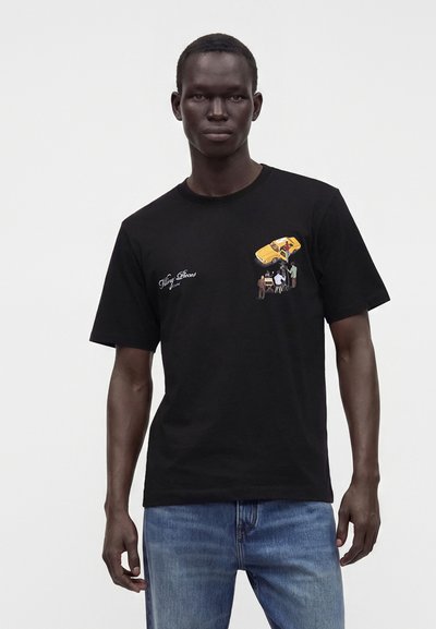 Άντρας που φοράει μαύρο T-shirt με μικρό γραφικό ανθρώπων που σηκώνουν ένα κίτρινο αυτοκίνητο και μπλε τζιν σε απλό φόντο.