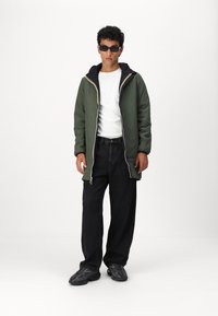 Veste isolée verte avec capuche noire et fermeture éclair contrastante, portée sur un t-shirt blanc, assortie à un pantalon noir ample et des chaussures noires texturées.