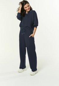 Frau lächelt, trägt einen navyblauen Sweatshirt mit Kragen und passende Jogginghose mit weißen Sneakers, Hand in der Tasche, posiert vor einem weißen Hintergrund.