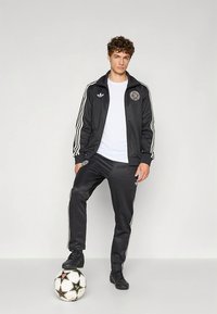 Juoda sportinė apranga su baltomis juostelėmis, sudaryta iš užtrauktuku užsegamos striukės ir kelnių. Dėvima ant balto marškinėlio, modelis stovi ant futbolo kamuolio.