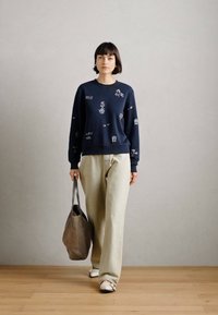 Σκούρο μπλε φούτερ με κεντήματα, συνδυασμένο με χαλαρούς μπεζ παντελόνες και λευκά αθλητικά παπούτσια. Κρατάει μια υφασμάτινη γκρι τσάντα.