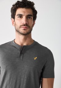 Grijs shirt met korte mouwen, ronde halslijn en een drieknopenlijst. Heeft een klein geel vogel logo op de borst. Van een katoenmix.
